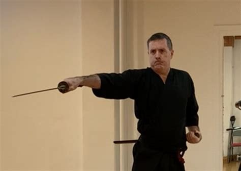 Iaidō practitioner in black training clothes extends a drawn sword to one side.
