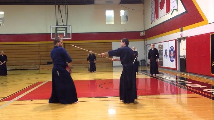 Jōdō partners demonstrate a staff technique in a gymnasium while other practitioners watch.
